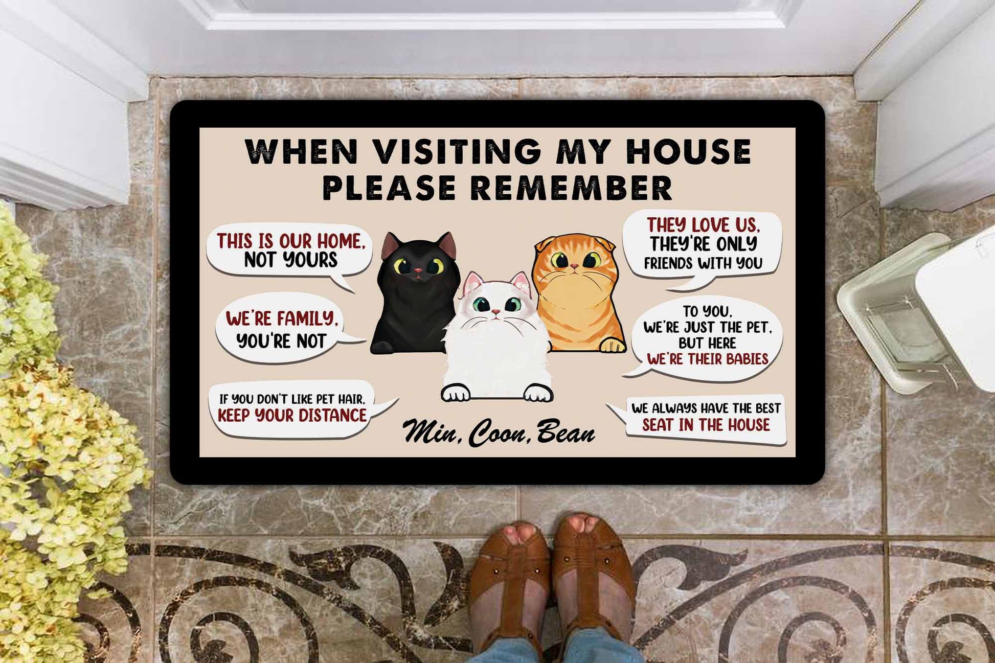 Doormat with cartoon cats and humorous text message on a tiled floor.