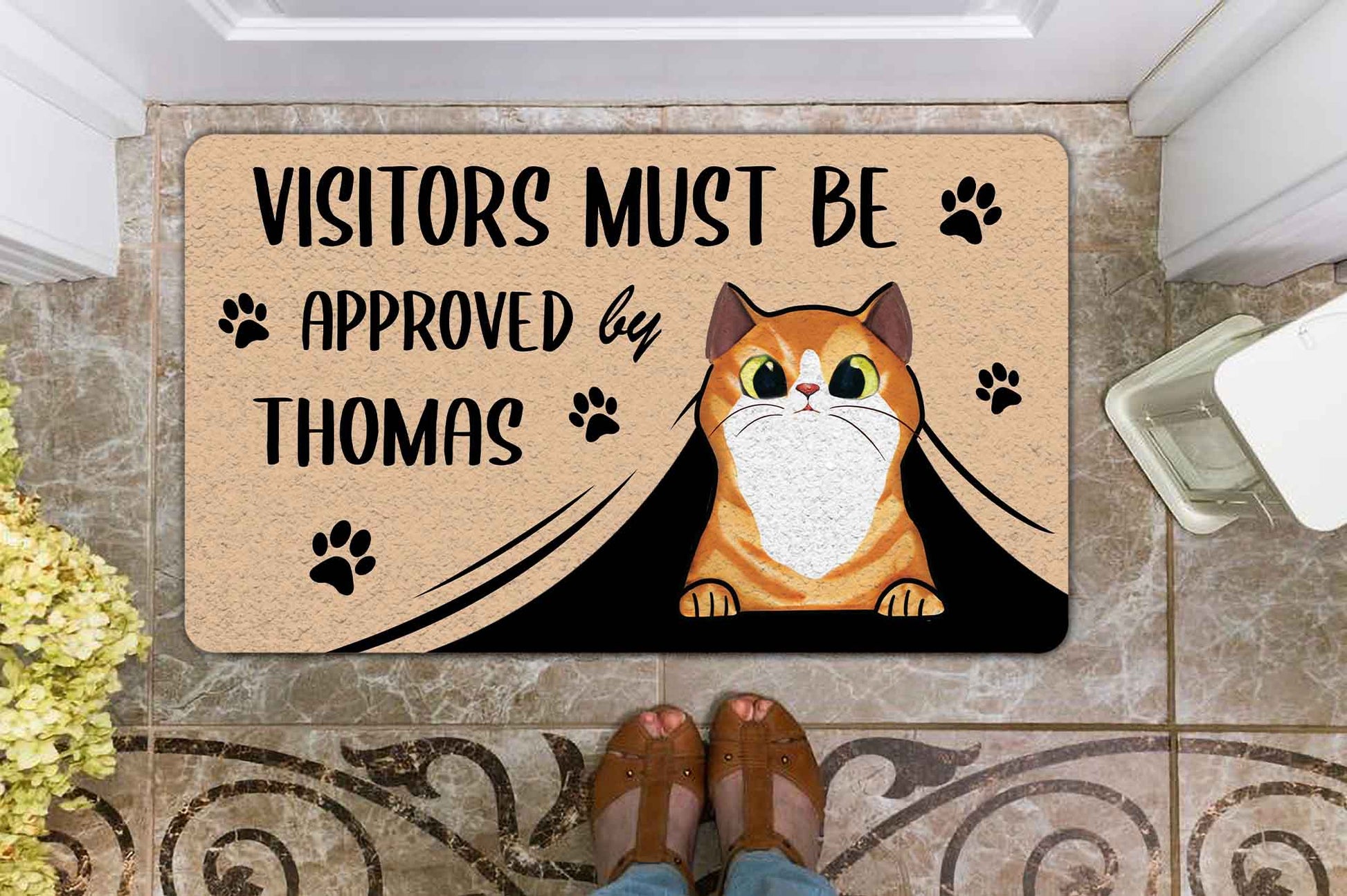 Doormat with a cat illustration and text 'Visitors must be approved by Thomas' on a tiled floor.