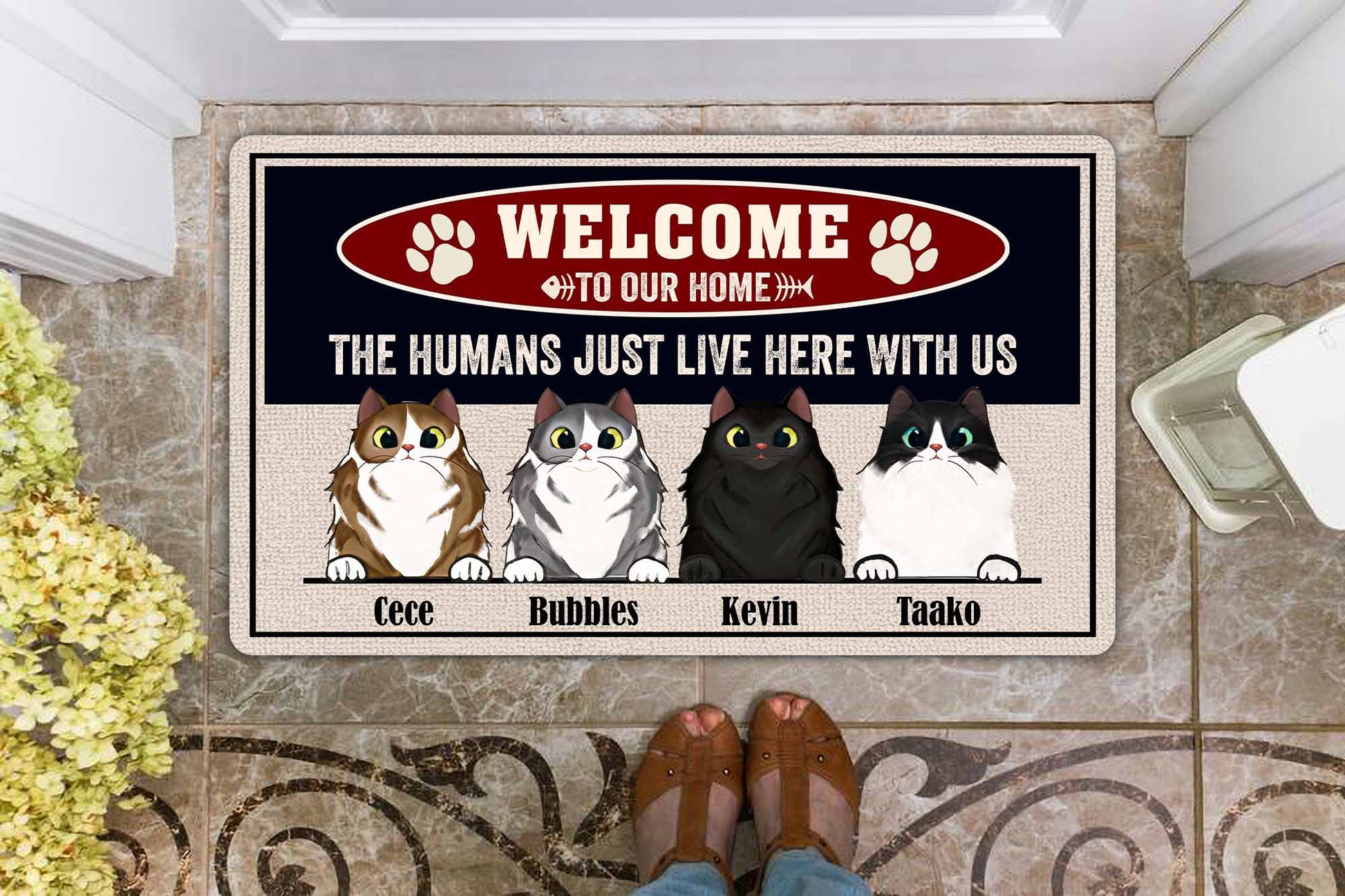 Doormat with cartoon cats and text 'Welcome to our home, the humans just live here with us' on a tiled floor.