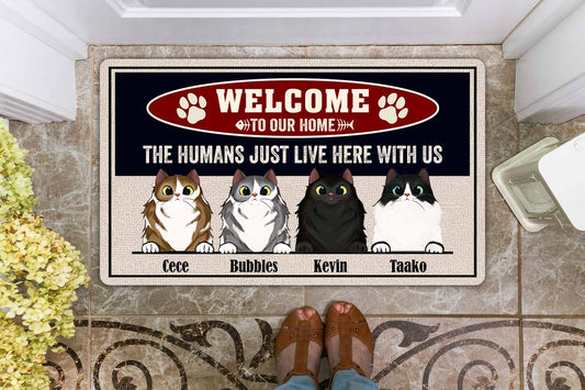 Doormat with cartoon cats and text 'Welcome to our home, the humans just live here with us' on a tiled floor.